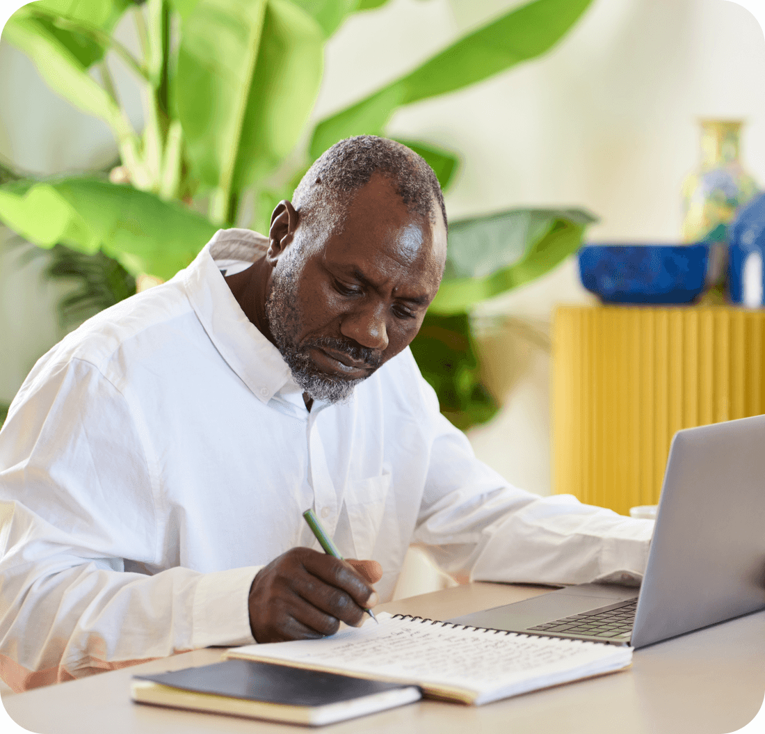 A photo of a man sat at a desk in front of a laptop and writing on a notepad