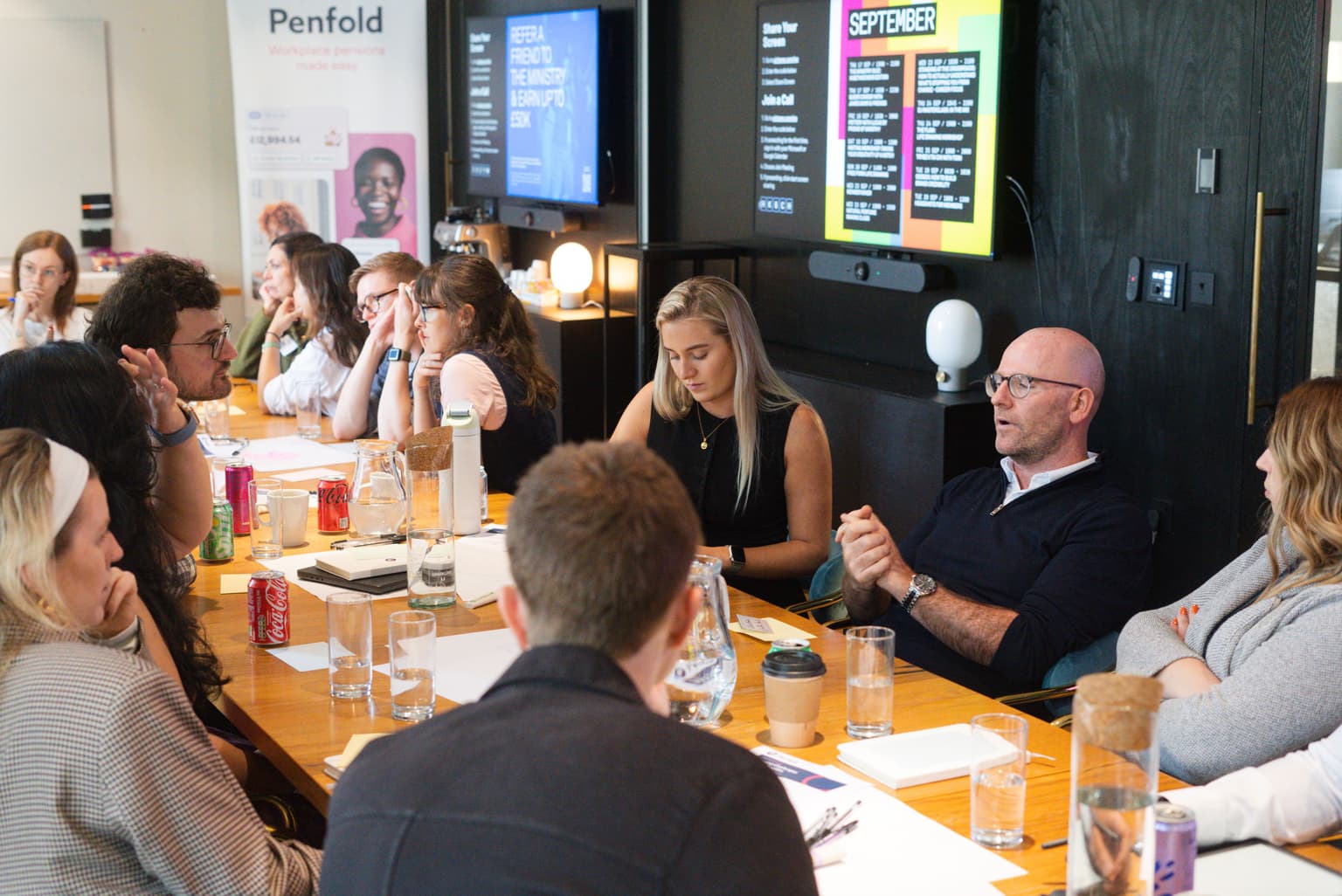 A group of people sit around a long table in a modern meeting room, with one man speaking and gesturing.